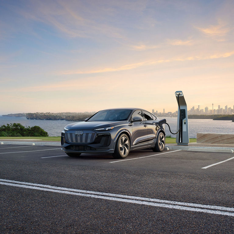 Three-quarter front view of a grey Audi Q6 Sportback e-tron parked and charging by a waterfront.