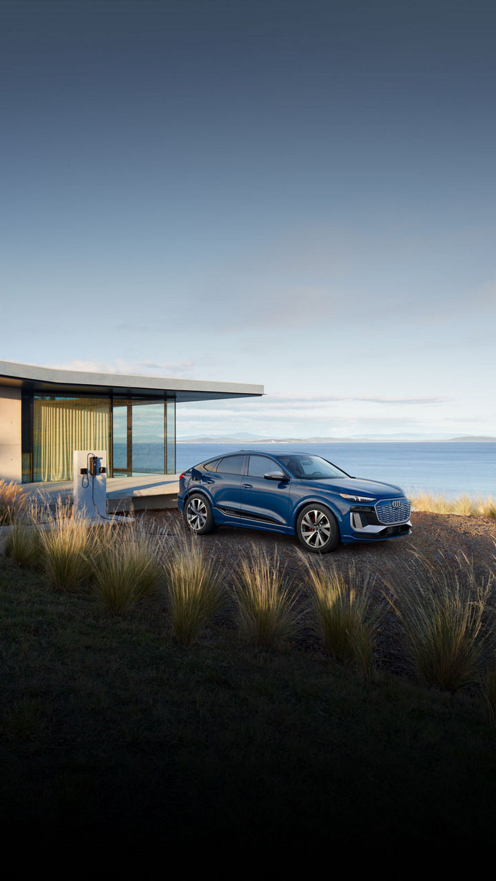 Three-quarter front view of a blue Audi SQ6 Sportback e-tron parked and charging by a waterfront home.