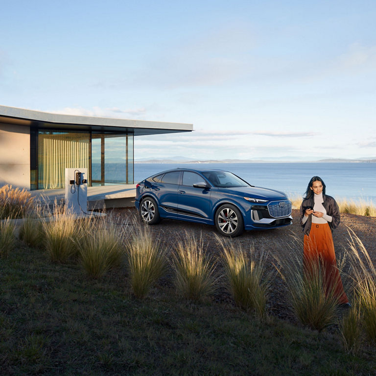 Side view of a blue Audi SQ6 Sportback e-tron parked and charging by a waterfront home with a person standing nearby.