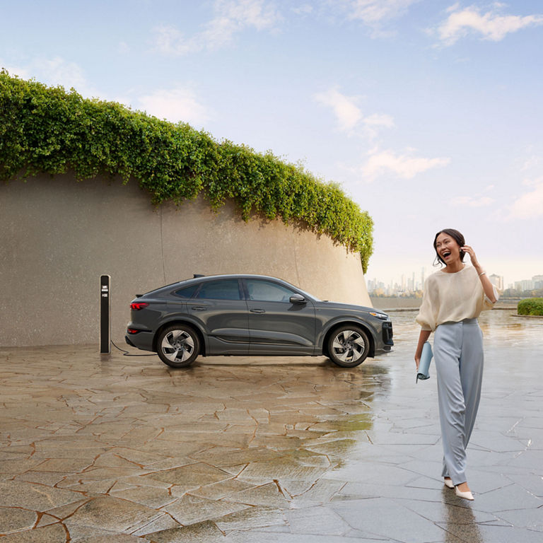 Vue latérale d'une Audi Q6 Sportback e-tron grise garée et en charge avec une personne marchant et riant à proximité.