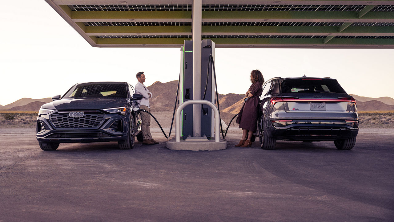 A man and a woman both charging their Audi Q8 e-trons at an Electrify America charging station. 