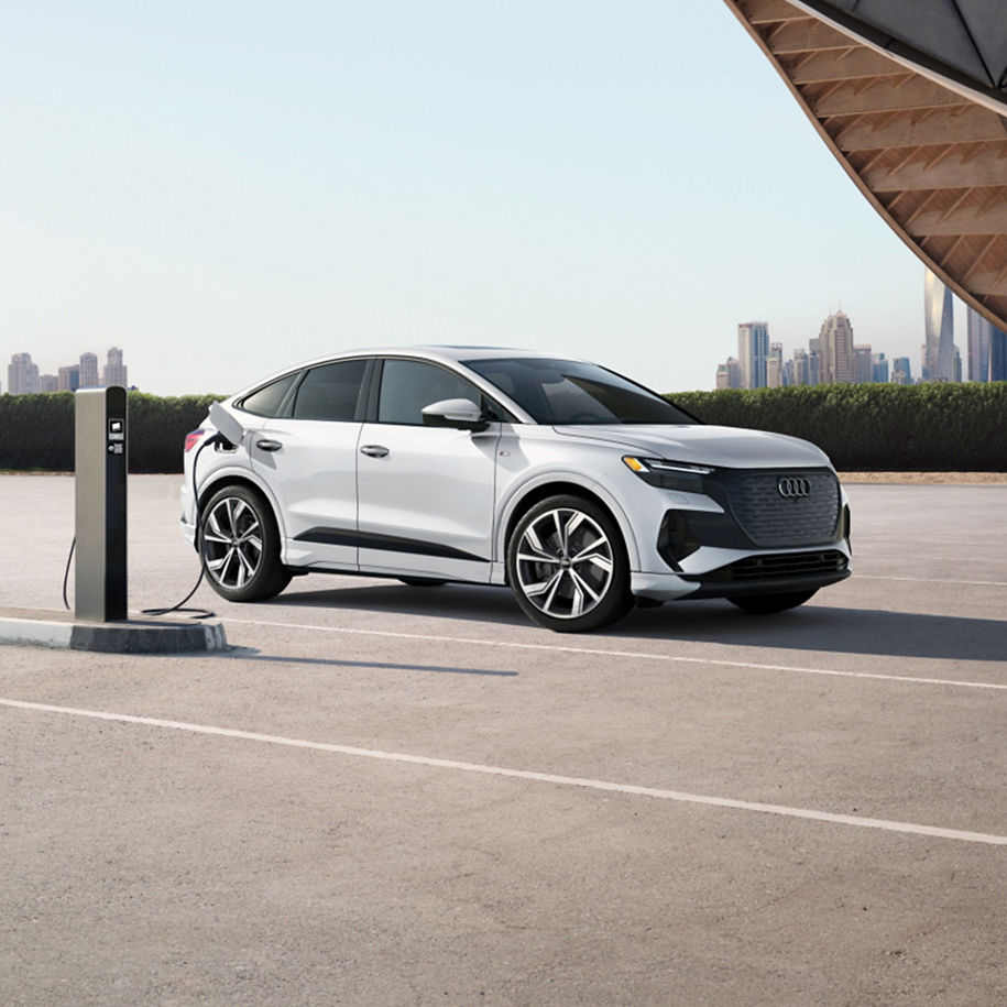 Three-quarter front view of a white Audi Q4 Sportback e-tron charging in front of city skyline. 