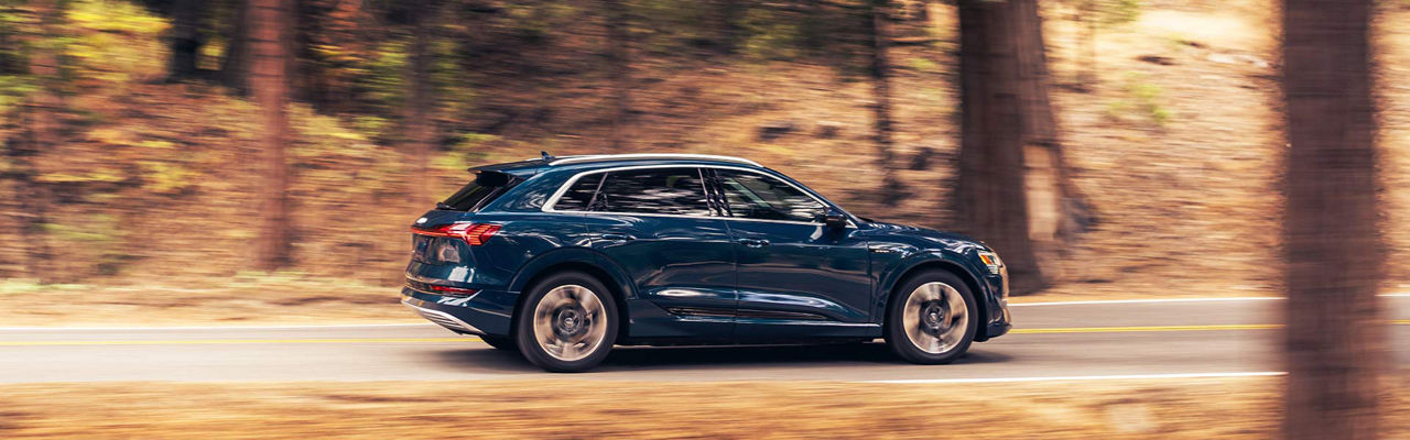 A blue Audi e-tron driving on the road, surrounded by trees and hills.