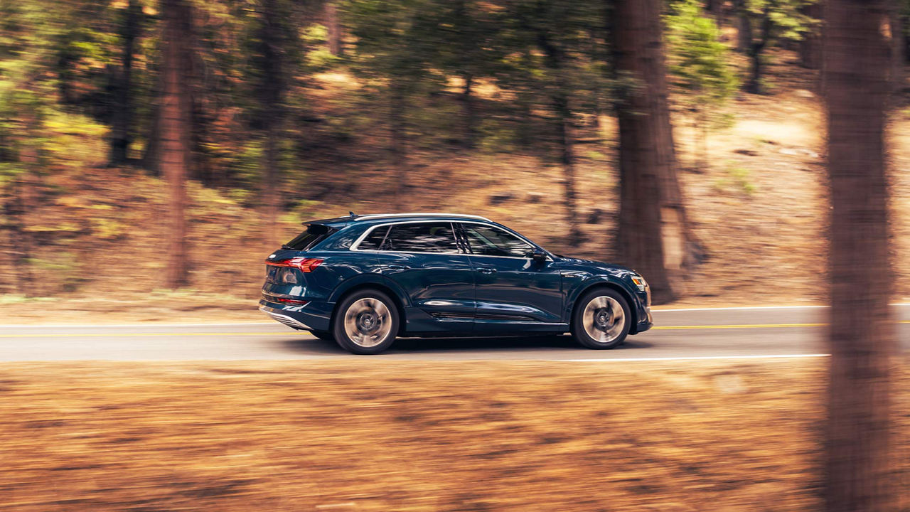 A blue Audi e-tron driving on the road, surrounded by trees and hills.