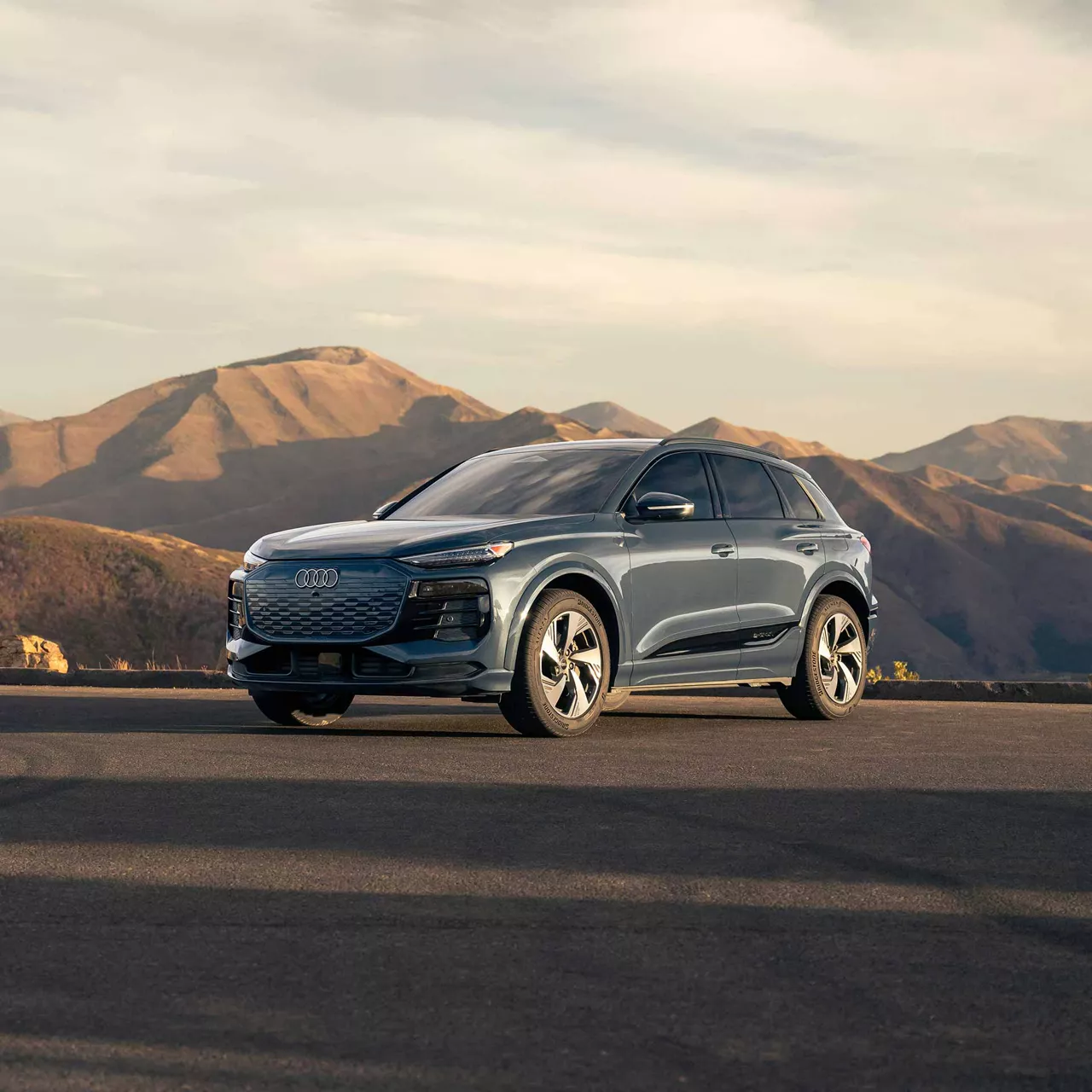 Three-quarter front view of a gray Audi A7 Sportback with illuminated headlights in front of a sunset. 