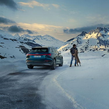 A wide-shot photo of a man in a parka and his dog near a parked Audi e-tron with snow-covered mountains in the background.