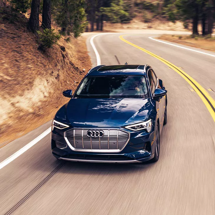 A mid-shot photo of an Audi e-tron driving down a curvy road in a forest.