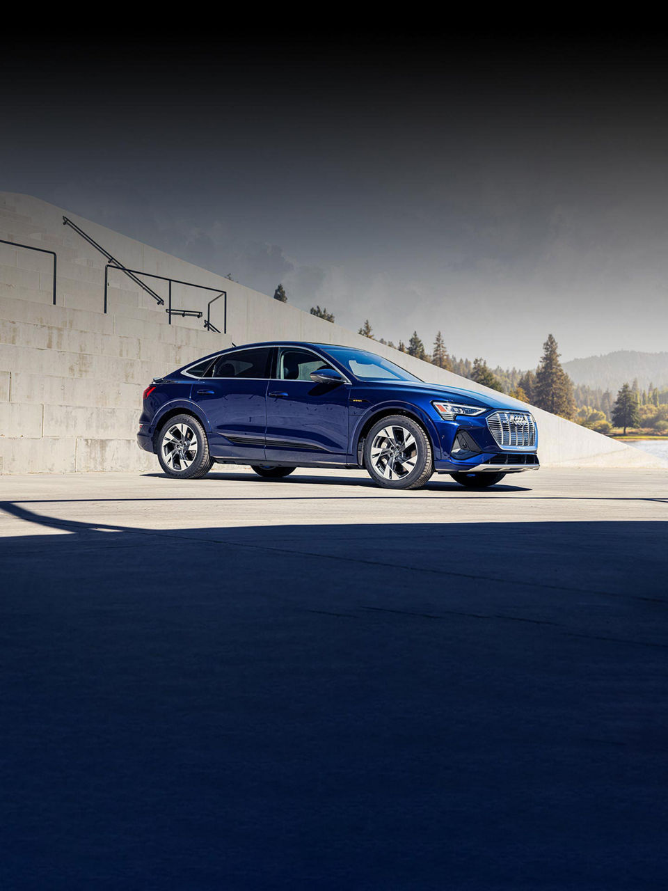 A mid-shot photo of an Audi e-tron parked in a parking lot with fir trees in the distance.