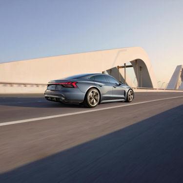 A mid-shot photo of an Audi e-tron GT driving down a highway past a modern-looking bridge.