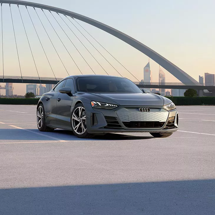 A wide-shot photo of an Audi e-tron parked in a concrete parking lot near a modern-looking bridge with a city skyline in the background.