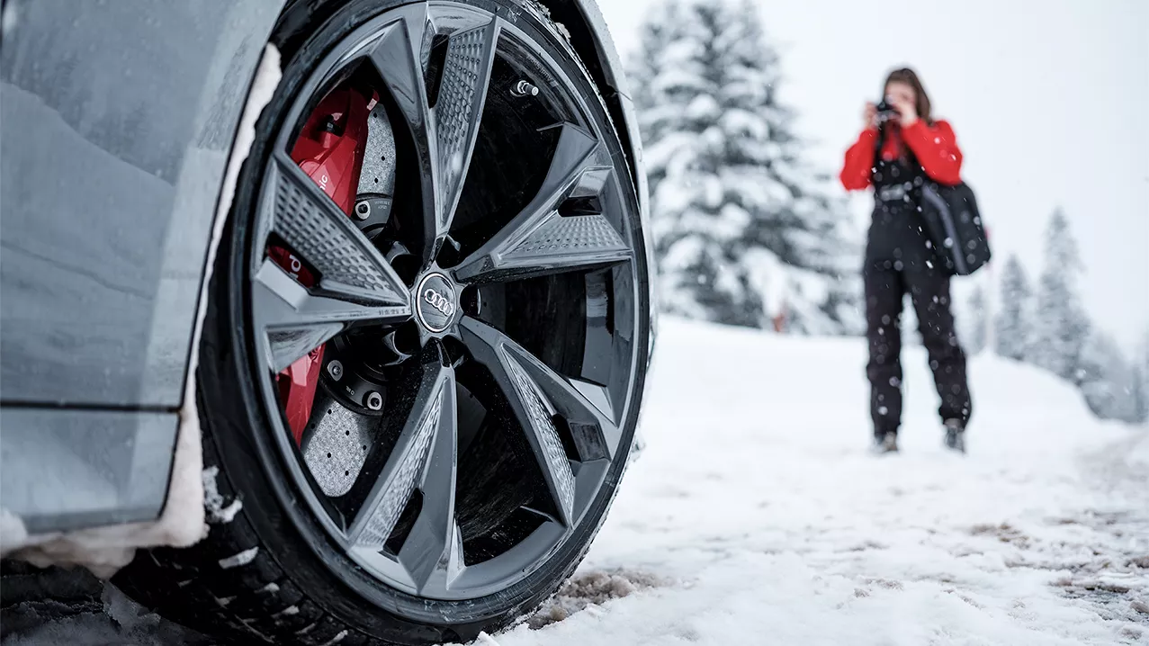 Close-up on the front wheel of an Audi vehicle in a winter landscape with a person standing in the background. 