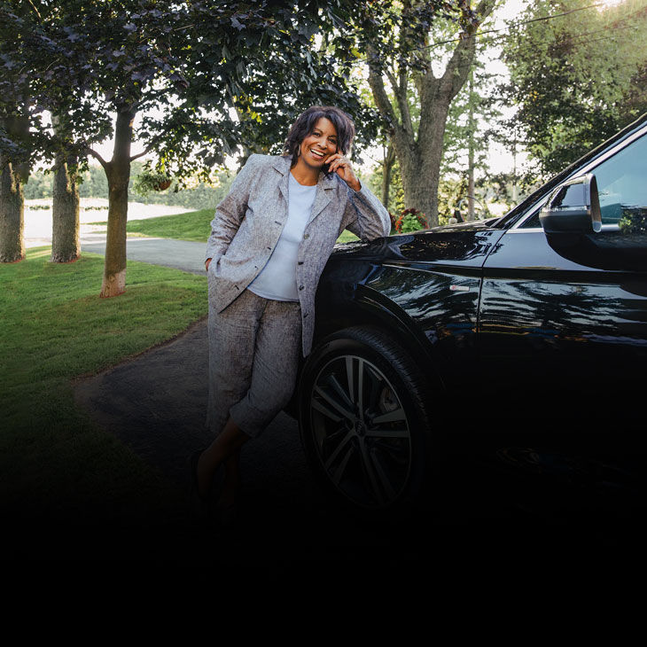 Front-profile of Nerene Virgin in a grey suit leaning on an black Audi vehicle. 