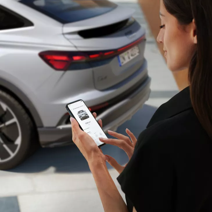 A woman’s hand gripping a smartphone opened to the myAudi app, while standing in front of an Audi e-tron.