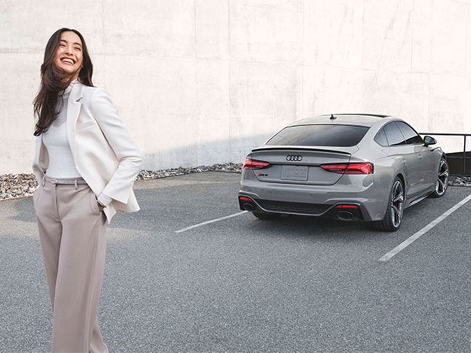 A person standing and laughing with an Audi RS 5 Sportback parked in the background.
