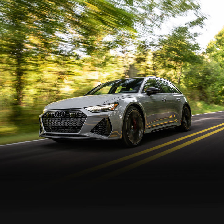 Three-quarter front view of a silver Audi RS 6 driving on a forested road. 