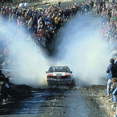  An early Audi motorsport vehicle driving through a large puddle, spraying the crowd on both sides. 