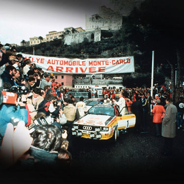  An early Audi motorsport vehicle at the finish line of the Monte Carlo Rally, surrounded by fans and photographers. 