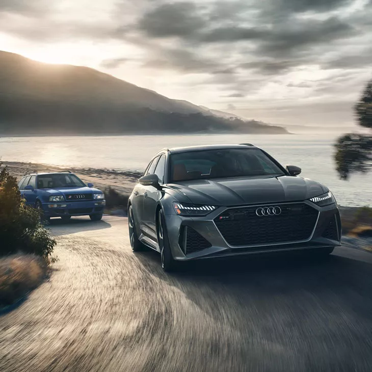 Two Audi Sport vehicles driving up a coastal road at sunset. 