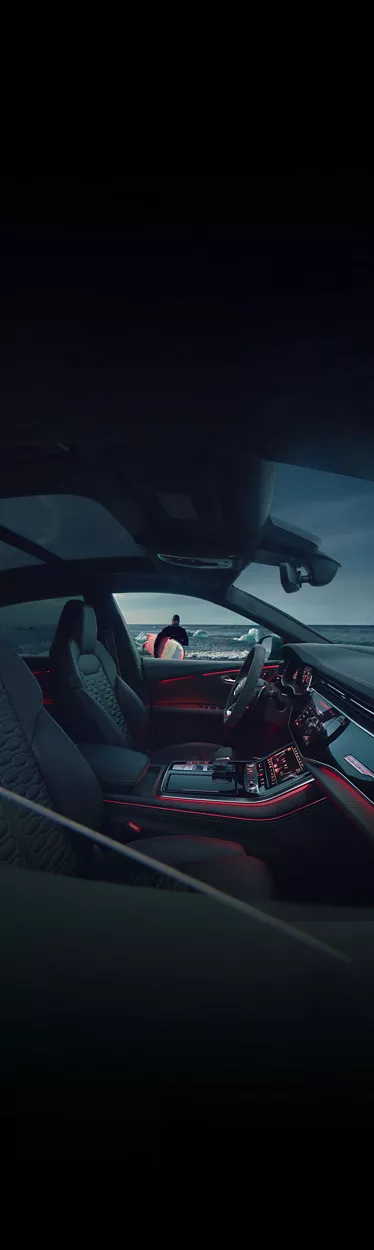 Interior shot of an Audi illuminated with red lights, as the owner approaches with his surfboard. 