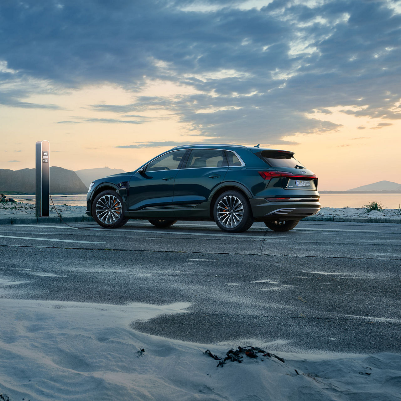 Profil latéral d’une Audi e-tron en recharge dans un stationnement près d’un plan d’eau.