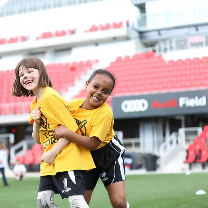 Two young girls in yellow DC SCORES soccer jerseys at a DC SCORES event on Audi Field.