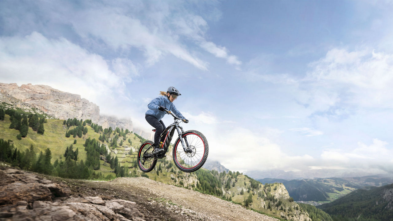 A woman performing a jump on her Audi electric mountain bike with a mountainous landscape behind her.