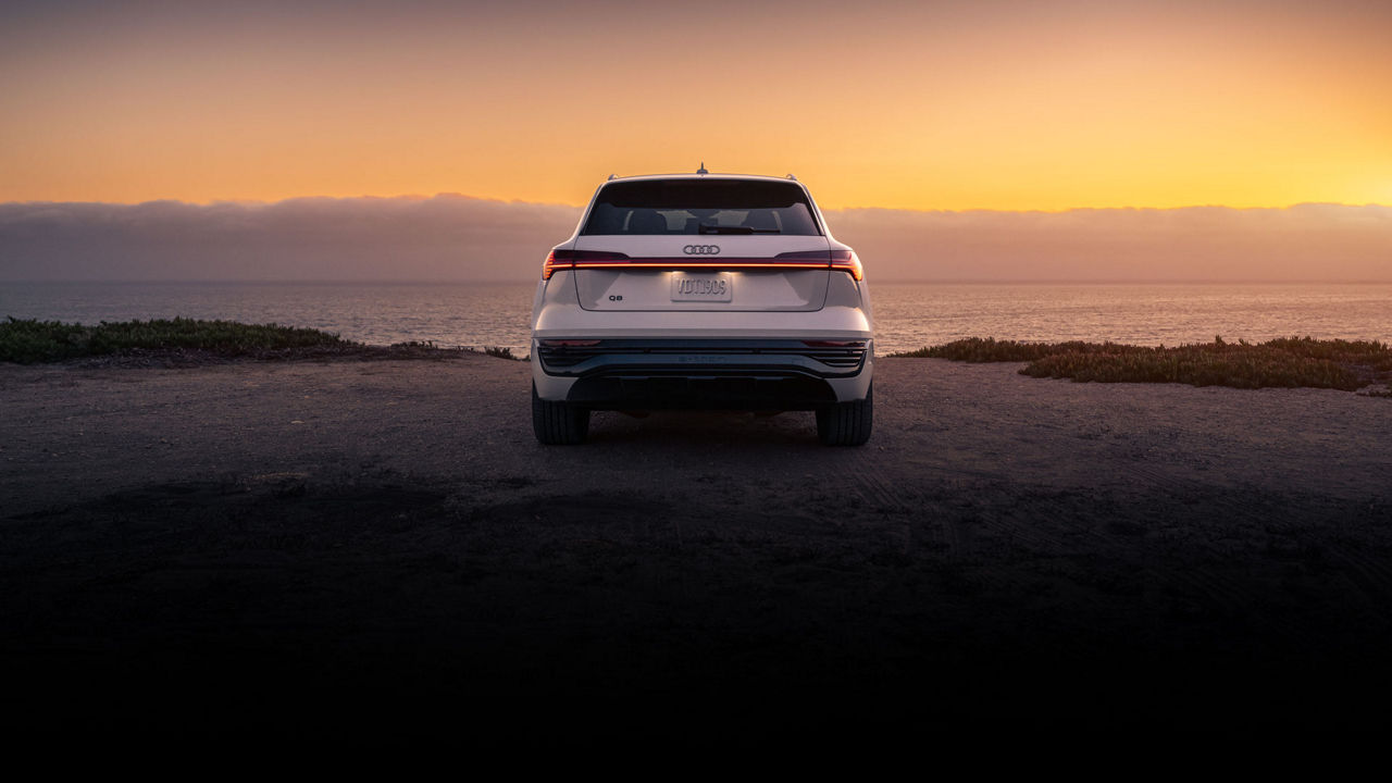 Rear view of a white Audi Q8 parked at the waterfront during sunset. 