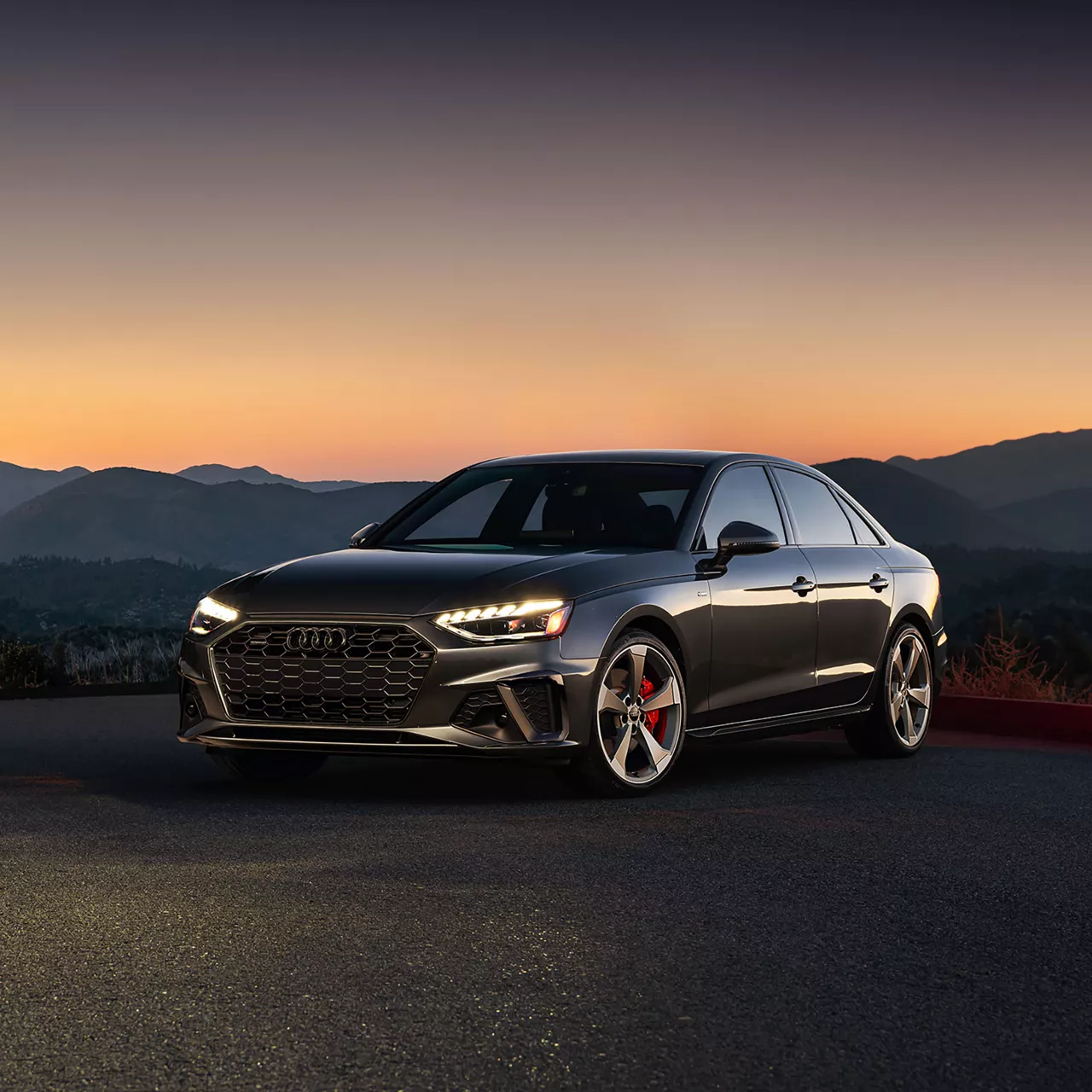 Three-quarter front view of a gray Audi A7 Sportback with illuminated headlights in front of a sunset. 