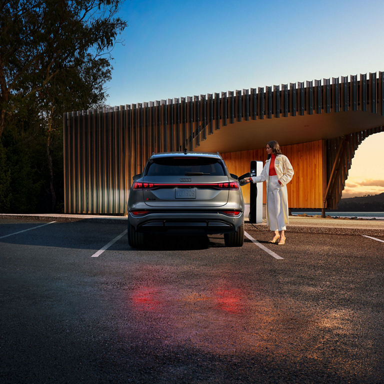 Rear profile of a person charging a grey Audi SQ6 e-tron by a modern building.