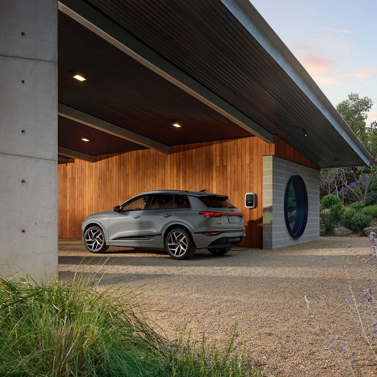 Three-quarter side view of a grey Audi SQ6 e-tron parked and charging at a modern house.