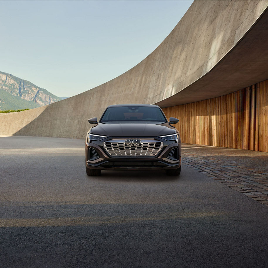 Vue avant d’un Audi Q8 Sportback e-tron brun stationné devant un bâtiment aux formes arrondies avec des montagnes en arrière-plan