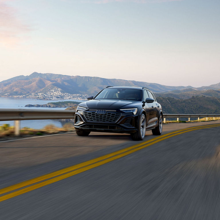 Elevated front-view of the SQ8 e-tron accelerating on an oceanside mountain road with mountains in the background.