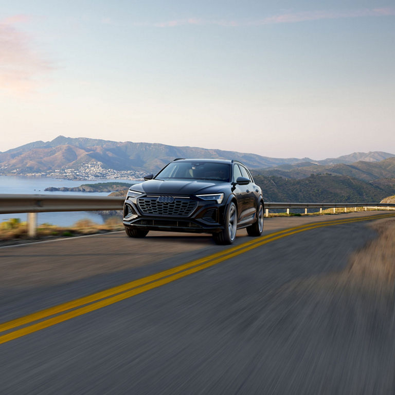 Elevated front-view of the SQ8 e-tron accelerating on an oceanside mountain road with mountains in the background.