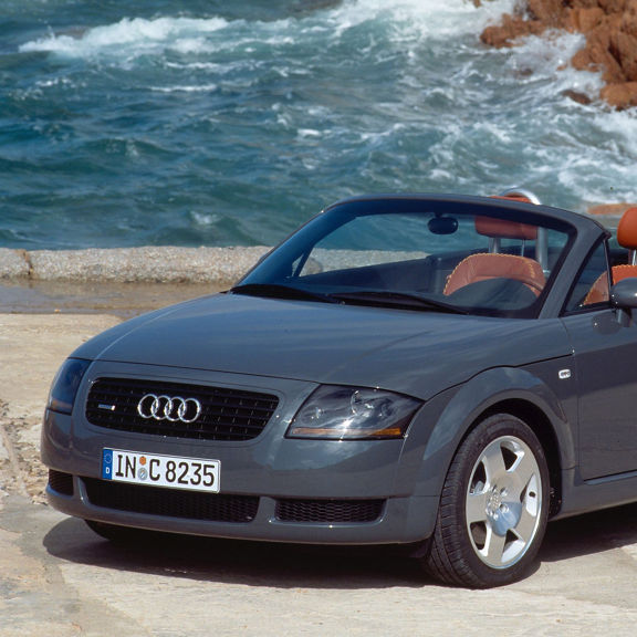 Front three-quarter view of a grey Audi TT Roadster parked next to a body of water.