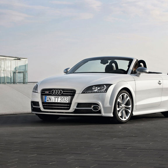 A white Audi TTS Coupe parked on a brick road next to a glass building.