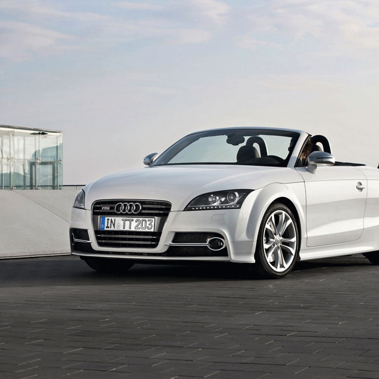 A white Audi TTS Coupe parked on a brick road next to a glass building. 