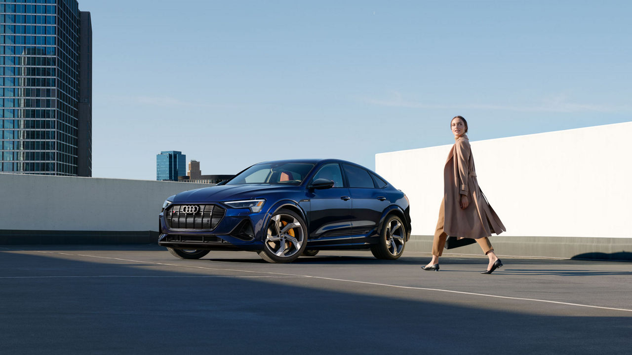 A woman walking towards an Audi e-tron in an empty parking lot with city buildings in the background.