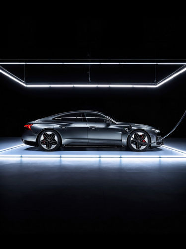 Shifting image of a gray Audi e-tron GT parked in front of a daytime city skline and on an illuminated platform in a dark room. 