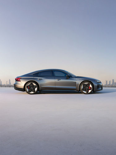 Shifting image of a grey Audi e-tron GT parked in front of a daytime city skline and on an illuminated platform in a dark room. 