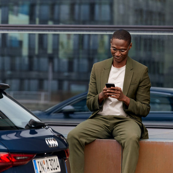 A man sitting next to his Audi while browsing the Discover tab on the myAudi app.