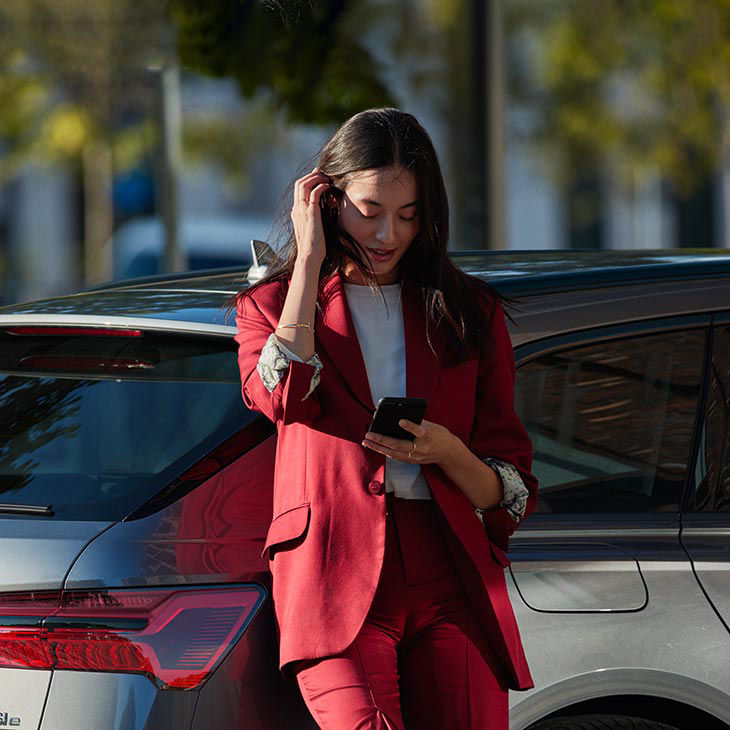 A mid-shot photo of a woman leaning against her Audi vehicle and looking down at her smartphone.