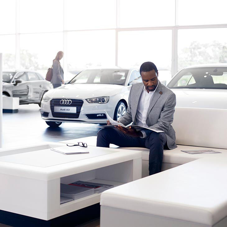 A far-shot photo of a man sitting in an Audi dealership reviewing his owner's manual.