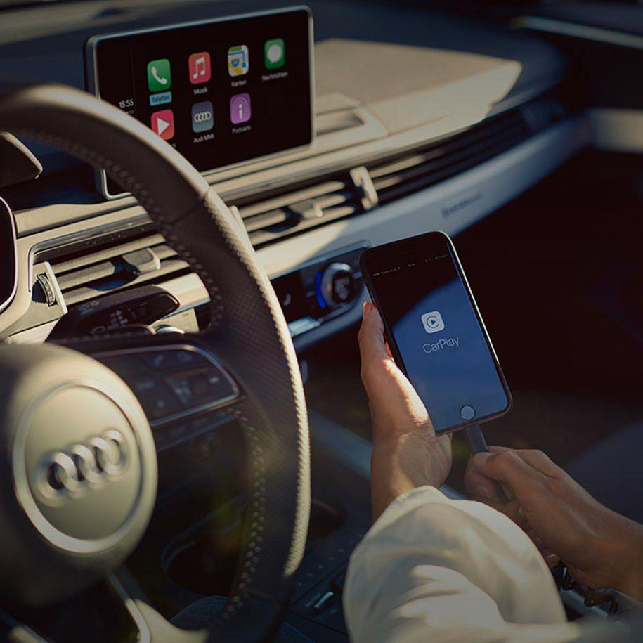 A close-up photo of a hand holding an Apple iPhone inside an Audi vehicle against a background of the Audi MMI technology system display.