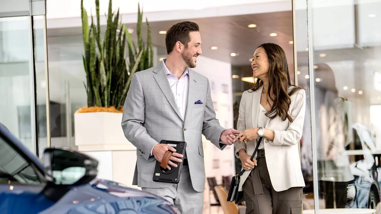 Audi sales specialist walking with a customer through the showroom. The sales specialist is handing the customer a key fob for an Audi vehicle. 