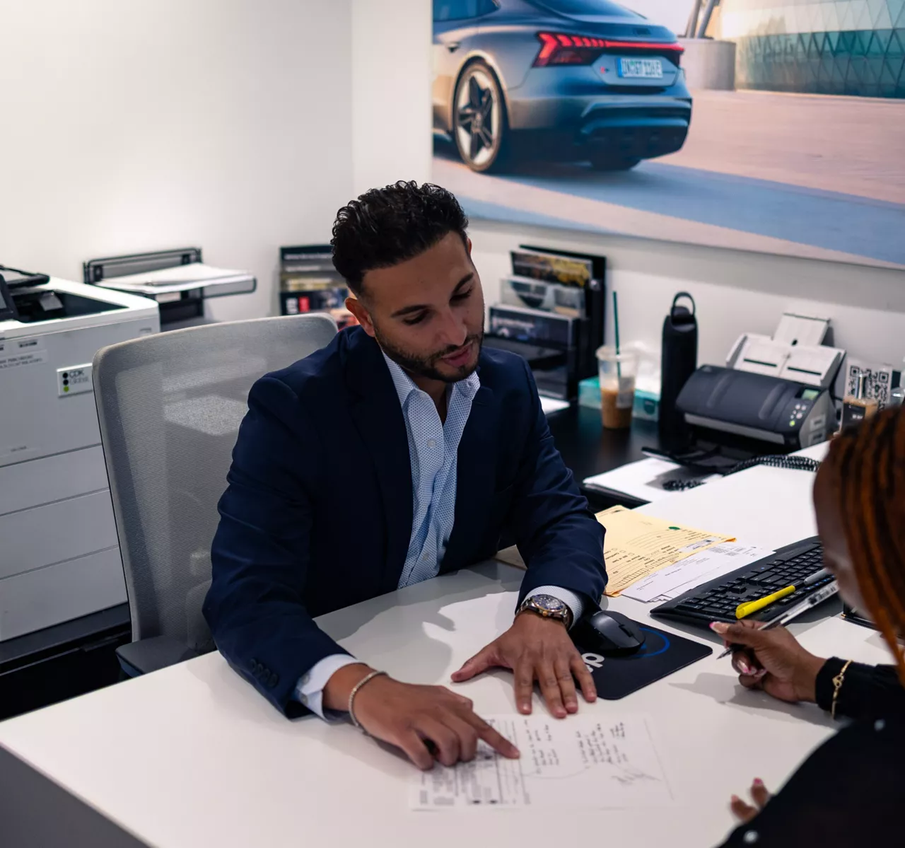Audi sales specialist walking with a customer through the showroom. The sales specialist is handing the customer a key fob for an Audi vehicle. 