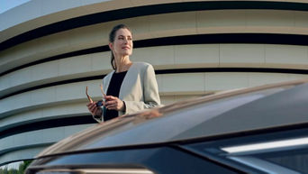 Woman holding glasses standing beside an Audi, looking to the right, smiling at the sun.