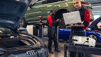 Audi technicians in a shop. One is working on a laptop, another one is working on brakes.
