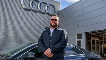 An Audi employee in front of an Audi vehicle and dealership building. 